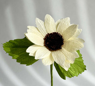 a row of pale buttercream yellow petals surrounds the dense black and brown center of this paper sunflower. Two olive green leaves complete the look and feel of this summertime staple.
