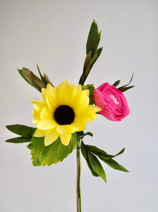 picture of bouquet featuring yellow paper sunflower with a black and brown center, bubble gum pink paper ranunculus and a sage green feather eucalyptus