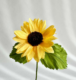 a row of bold yellow petals surrounds the dense black and brown center of this paper sunflower. Two olive green leaves complete the look and feel of this summertime staple.