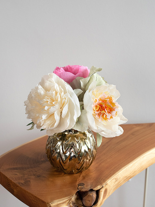 A handmade paper flower arrangement featuring 3 large, full-bloom peonies in shades of blush pink, ivory, and ivory with pink and cream interior. The delicate petals have realistic texture and gentle curves, arranged with sage green paper lamb's ear leaves. This arrangement has been placed in a decorative vase and sits on a wood table.