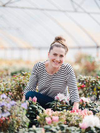 Smiling woman in a striped shirt sitting among colorful flowers in a greenhouse