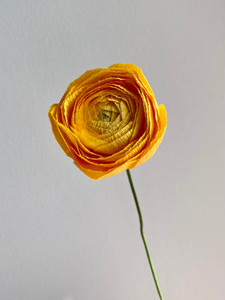 a handcrafted paper ranunculus with dozens of tightly layered, ruffled petals in shades of sunshine yellow, gradually opening from a dense center to a full bloom. The flower is attached to a slender green stem, capturing the delicate and intricate structure of a real ranunculus