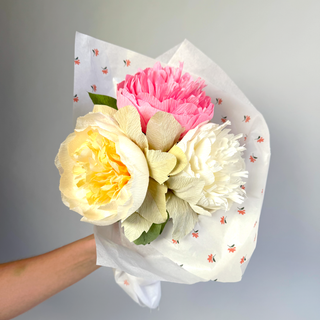 Bouquet of paper peonies wrapped in patterned paper held by a hand against a plain background