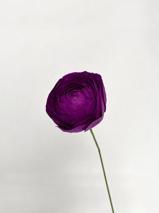 a handcrafted paper ranunculus with dozens of tightly layered, ruffled petals in shades of dark purple, gradually opening from a dense center to a full bloom. The flower is attached to a slender green stem, capturing the delicate and intricate structure of a real ranunculus