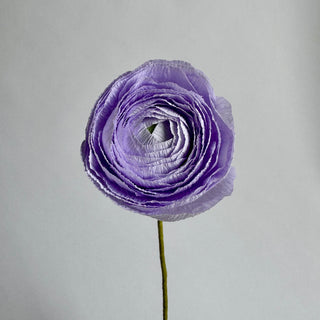 a handcrafted paper ranunculus with dozens of tightly layered, ruffled petals in shades of lilac purple, gradually opening from a dense center to a full bloom. The flower is attached to a slender green stem, capturing the delicate and intricate structure of a real ranunculus