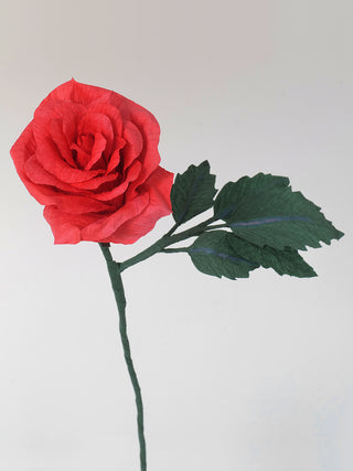 A handcrafted paper rose with delicately layered petals in bright shades of coral, curled and shaped to mimic the natural texture of a real rose. The flower is attached to a slender green stem with realistic-looking leaves made from emerald green crepe paper.