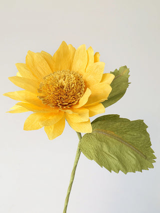 a row of bold yellow petals surrounds the dense yellow center of this paper sunflower. Two olive green leaves complete the look and feel of this summertime staple.