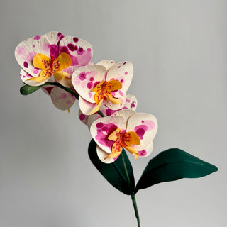 A single cardstock papaer orchid stem, featuring white petals with pink and magenta spots, yellow centers, and dark green leaves, set against a plain light gray background.