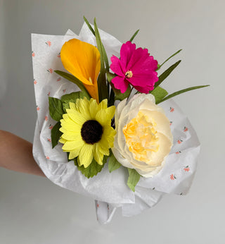 Bouquet of handcrafted Italian crepe paper flowers including an orange calla lily, hot pink cosmo, bright yellow sunflower and ivory and cream fluffy peony. The bouquet is accented with paper lemon leaves and paper lily grass; both bright green. The bouquet is wrapped and comes presented in white tissue paper with a floral pattern on a gray background