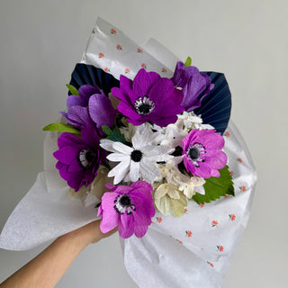 Bouquet of purple, white, and pink flowers wrapped in white paper with a floral pattern on a plain background.