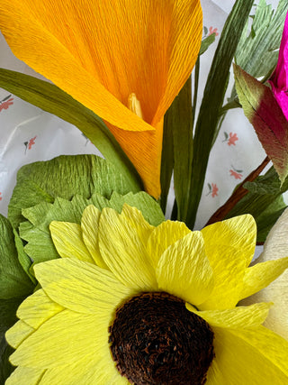 Close-up of paper flowers including a yellow sunflower and orange calla lily on a patterned background.
