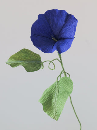 A delicate handmade paper flower resembling a morning glory with dark blue crepe paper petals, a pale yellow center, and green textured leaves. The flower is attached to a slender green stem with a curling tendril, set against a plain light gray background